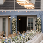 Long banquet table with blue and white floral centerpieces, candles, and crystal glassware under modern geometric chandelier
