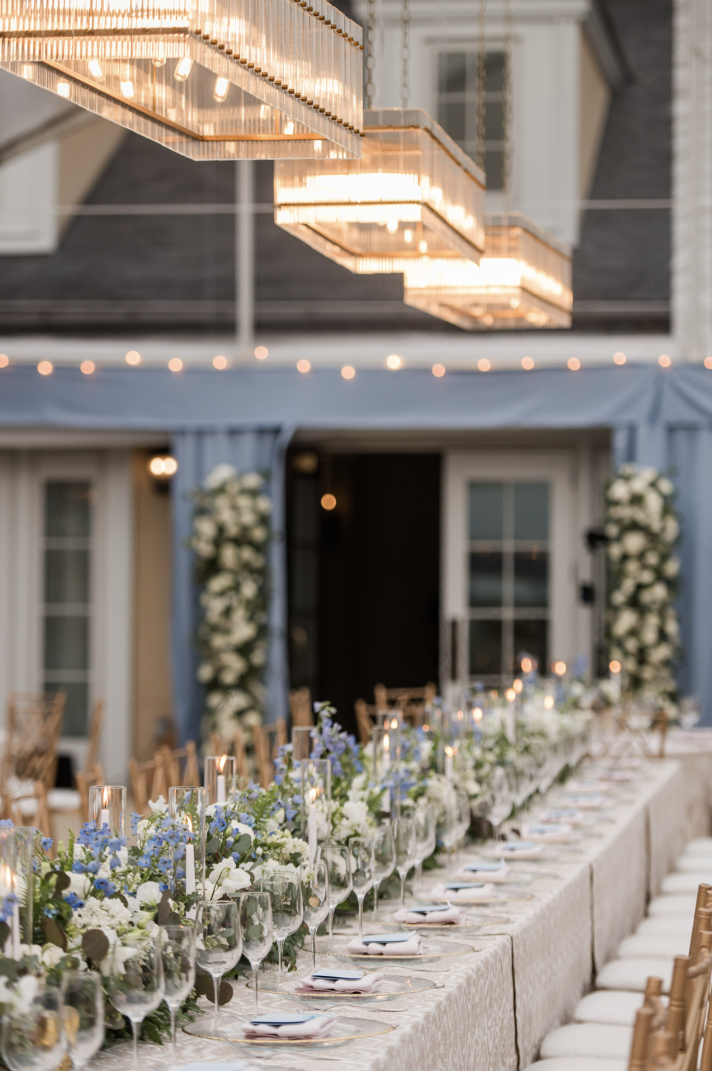 Long banquet table with blue and white floral centerpieces, candles, and crystal glassware under modern geometric chandelier