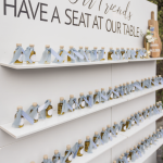 White shelving display with rows of small olive oil bottles tied with blue ribbons and sign reading "Olive Our Friends Have a Seat at Our Table"