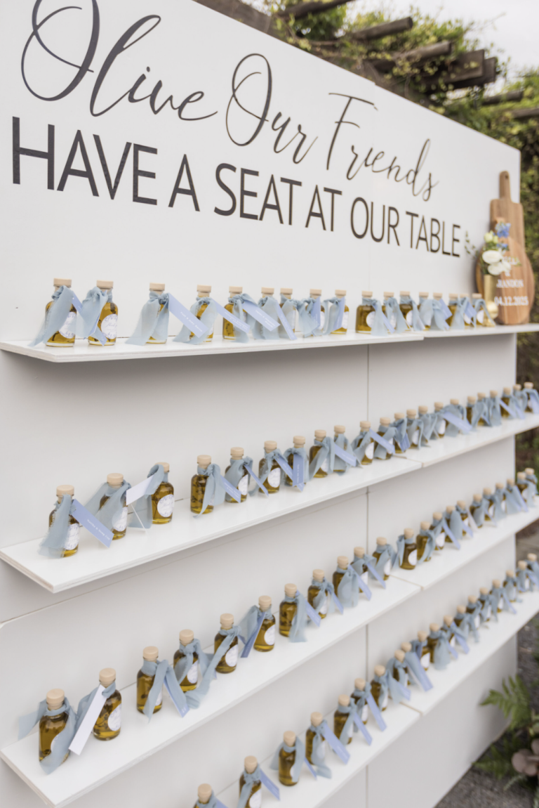 White shelving display with rows of small olive oil bottles tied with blue ribbons and sign reading "Olive Our Friends Have a Seat at Our Table"
