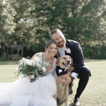 Newlyweds sitting on grass with their golden retriever in formal attire, bride holding white bouquet in garden setting