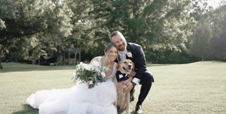 Newlyweds sitting on grass with their golden retriever in formal attire, bride holding white bouquet in garden setting