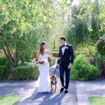 Newlywed couple with dog walking hand-in-hand through garden courtyard surrounded by lush greenery and white benches