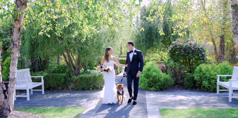 Newlywed couple with dog walking hand-in-hand through garden courtyard surrounded by lush greenery and white benches