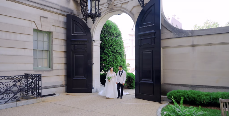 Couple walking through arched stone entrance of historic building with manicured greenery