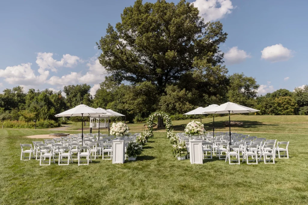 Outdoor wedding ceremony setup with white folding chairs, floral arch, and umbrellas under large tree on green lawn