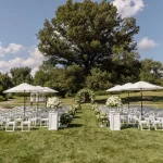 Outdoor wedding ceremony setup with white folding chairs, floral arch, and umbrellas under large tree on green lawn