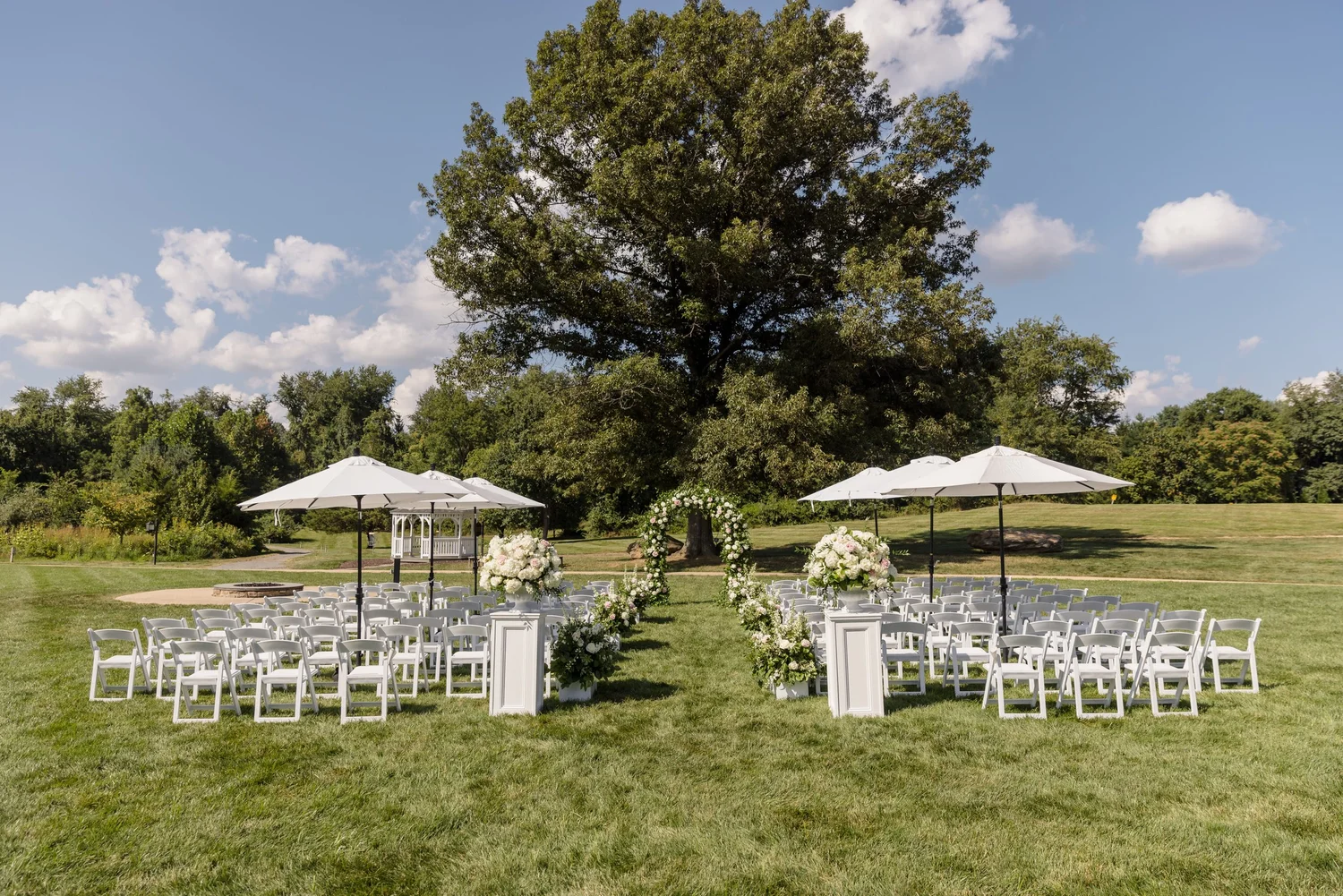 Outdoor wedding ceremony setup with white folding chairs, floral arch, and umbrellas under large tree on green lawn