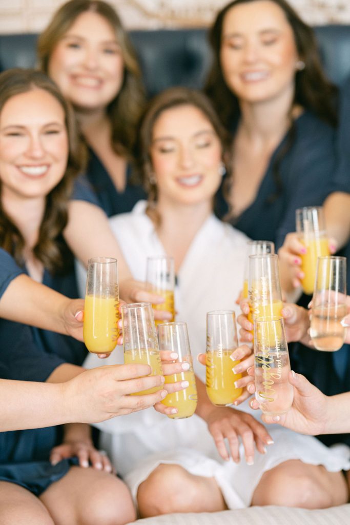 Bride and bridesmaids in navy robes toast with mimosas during wedding morning preparations