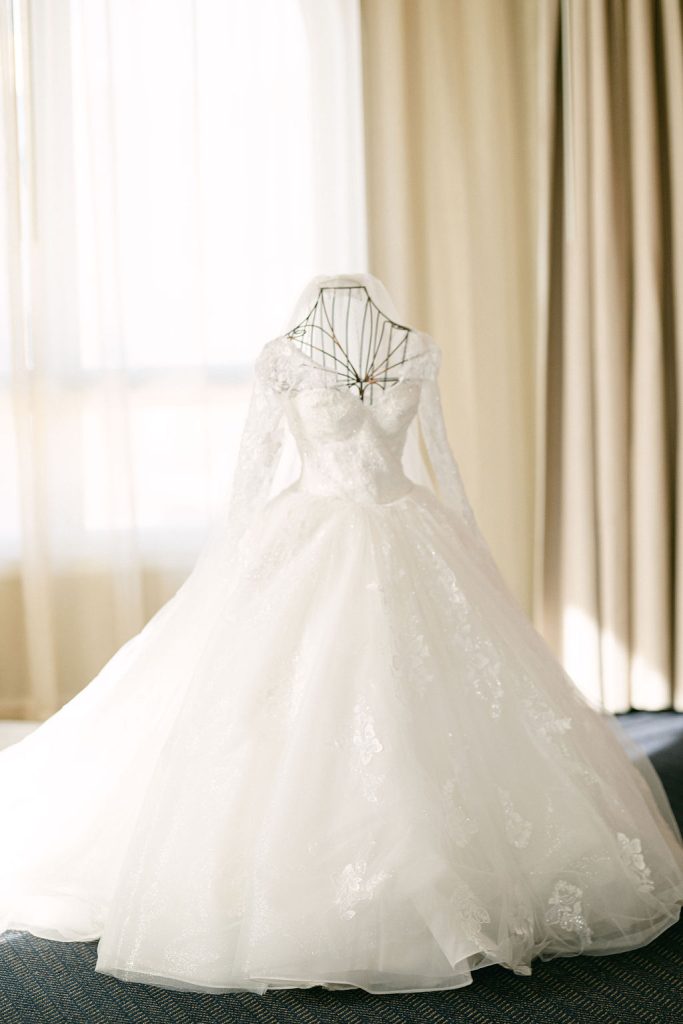 White lace ball gown wedding dress with long sleeves hanging on dress form near window