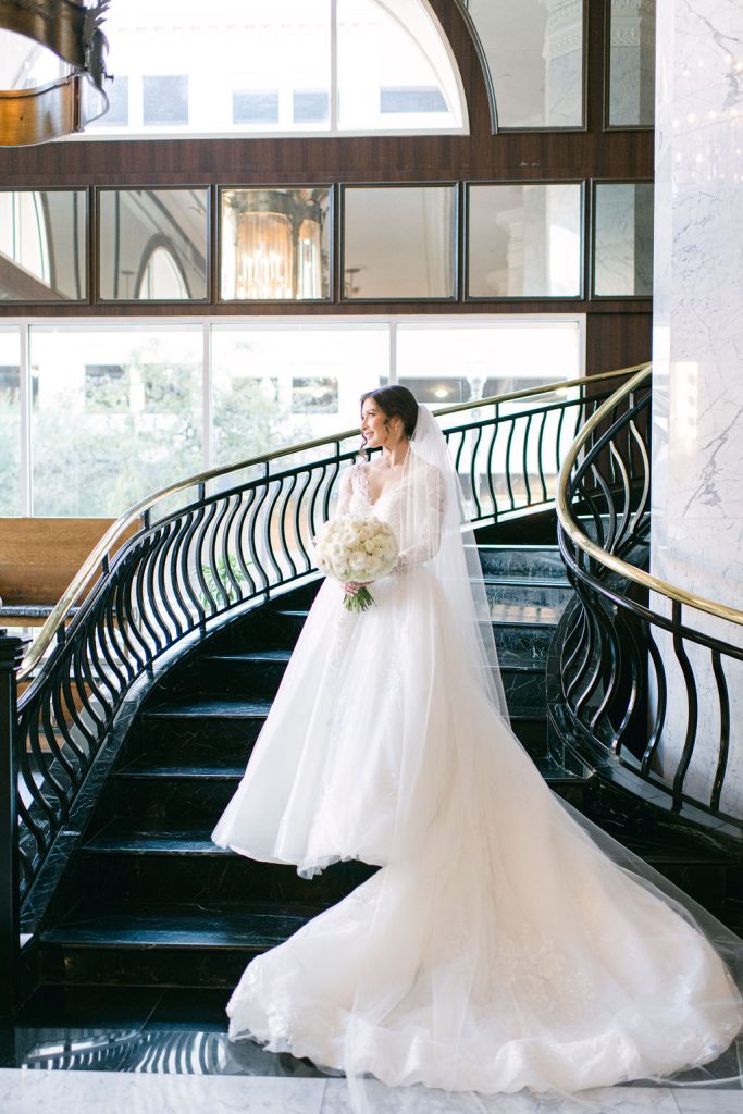 Bride Victoria posing on grand curved staircase in lace wedding gown with flowing cathedral train
