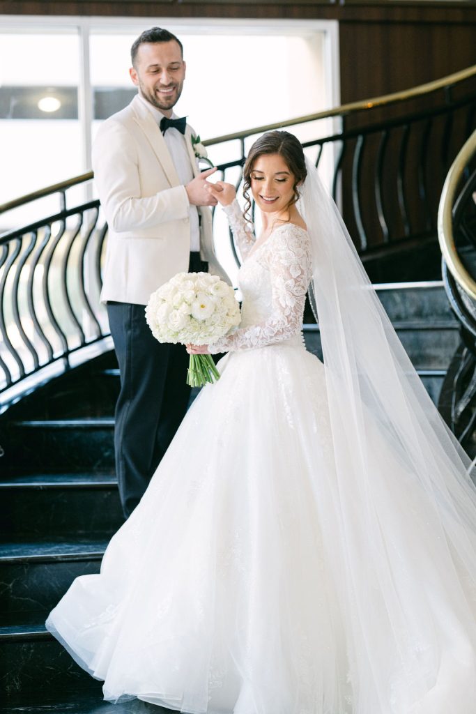 Victoria and Anthony descend grand staircase, bride in lace ballgown holding white rose bouquet