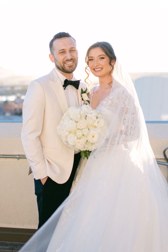 Victoria and Anthony pose together by the waterfront, bride holding white rose bouquet in lace gown with veil