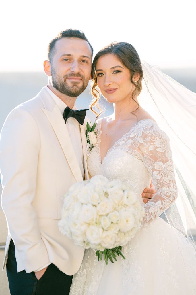 Victoria and Anthony standing together outdoors holding white rose bouquet with veil flowing in breeze