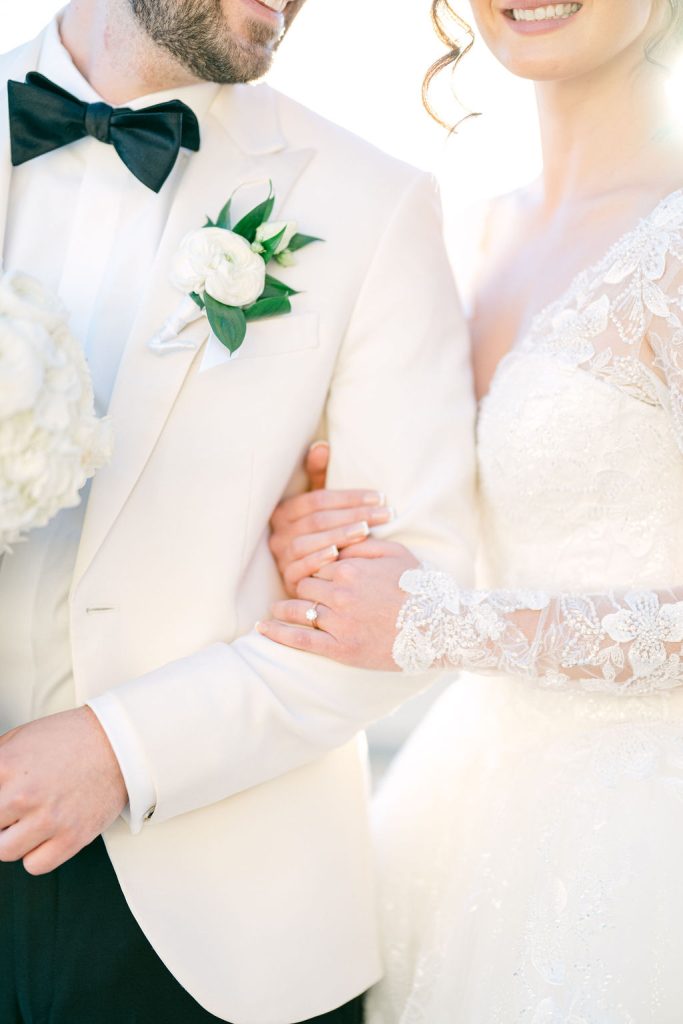 Groom in ivory tuxedo with emerald bow tie and white ranunculus boutonniere beside bride in lace gown