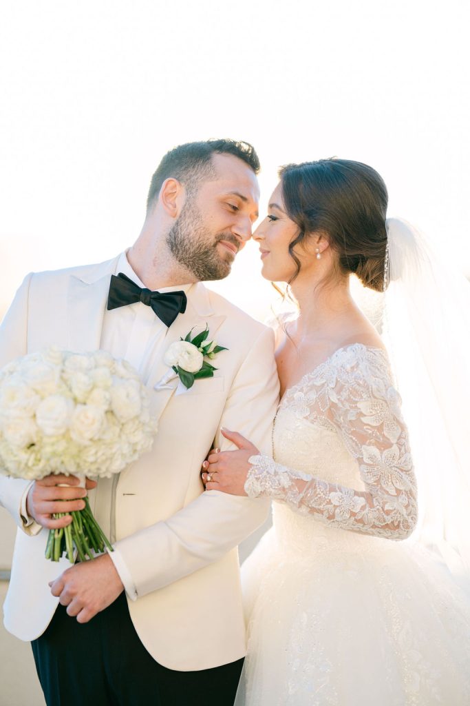 Victoria and Anthony embrace in their wedding attire, groom in ivory jacket with black bow tie, bride in lace gown holding white bouquet