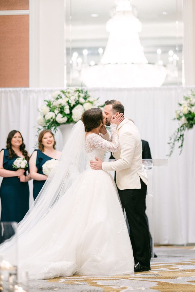 Victoria and Anthony share their first kiss as newlyweds beneath white floral arch with bridesmaids looking on