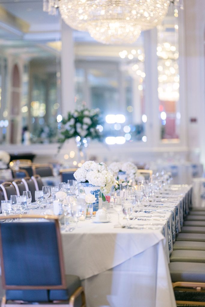 Elegant reception table with white linens, blue accents, white floral centerpieces, and crystal chandelier overhead