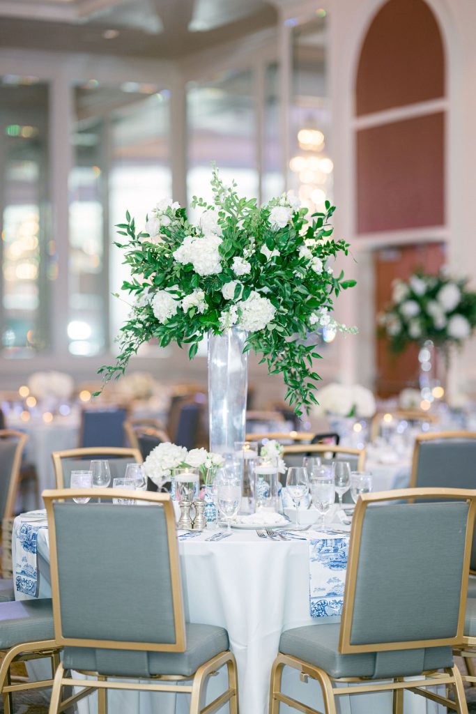 Tall white floral centerpiece with greenery on reception table set with blue and white china