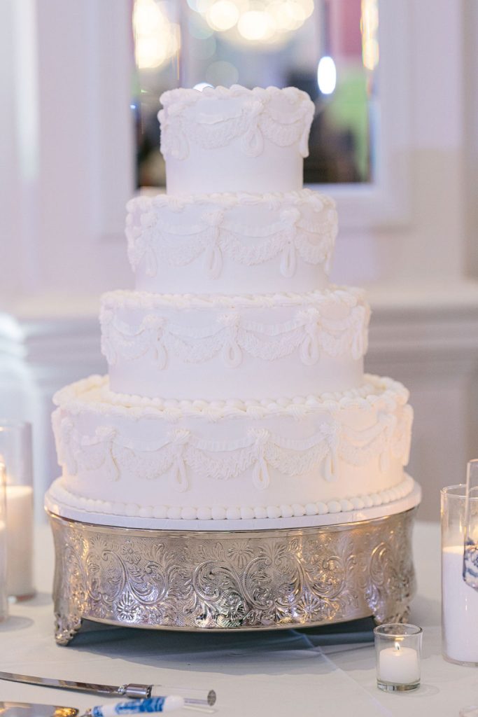 Four-tier white wedding cake with delicate lace piping on ornate silver pedestal stand