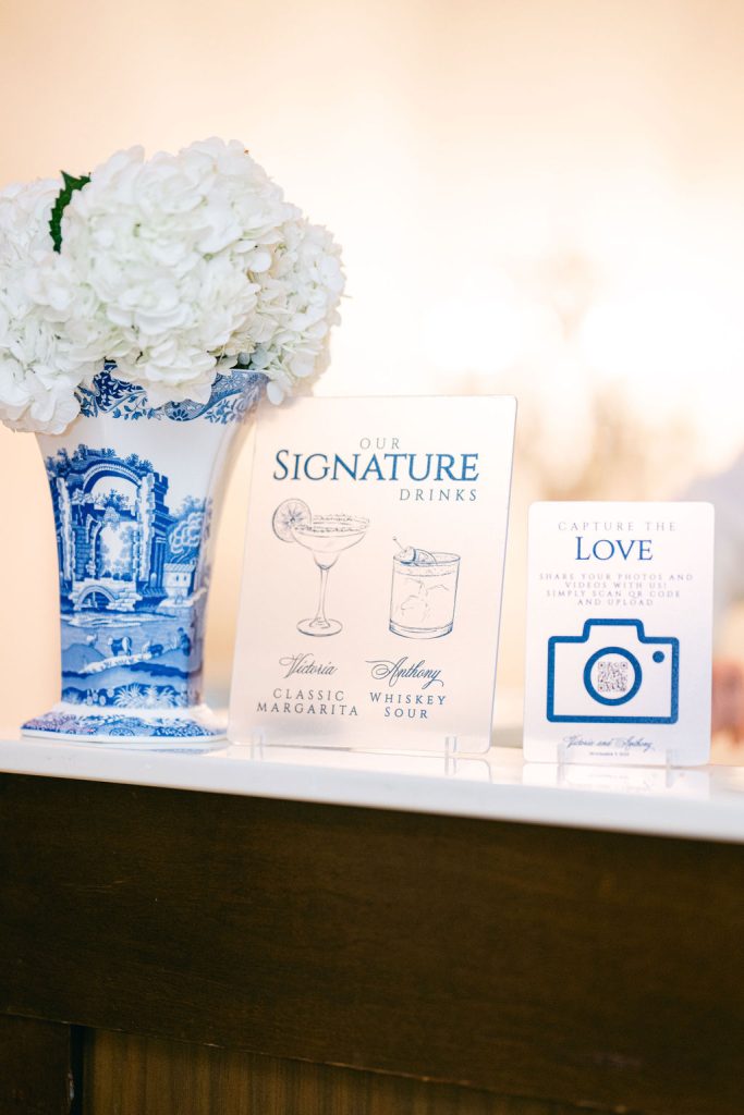 White hydrangea bouquet in blue-and-white porcelain vase beside signature drinks menu and social media hashtag sign