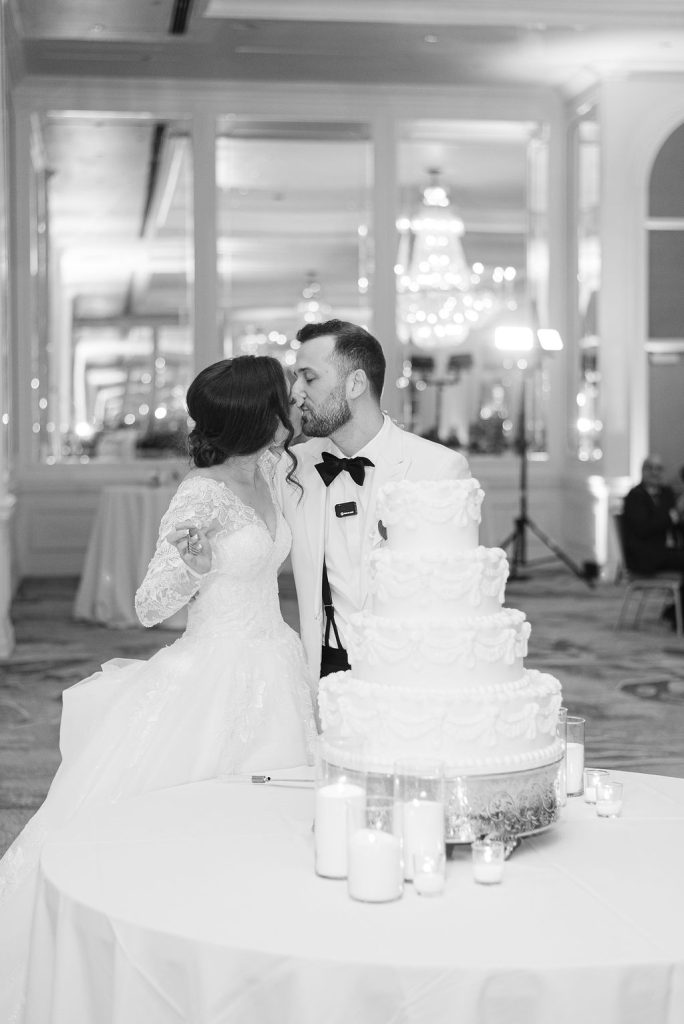 Victoria and Anthony kiss behind their tiered white wedding cake surrounded by candles in an elegant ballroom
