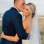 Bride with elegant updo and veil smiles at camera while groom in navy suit kisses her temple at outdoor venue