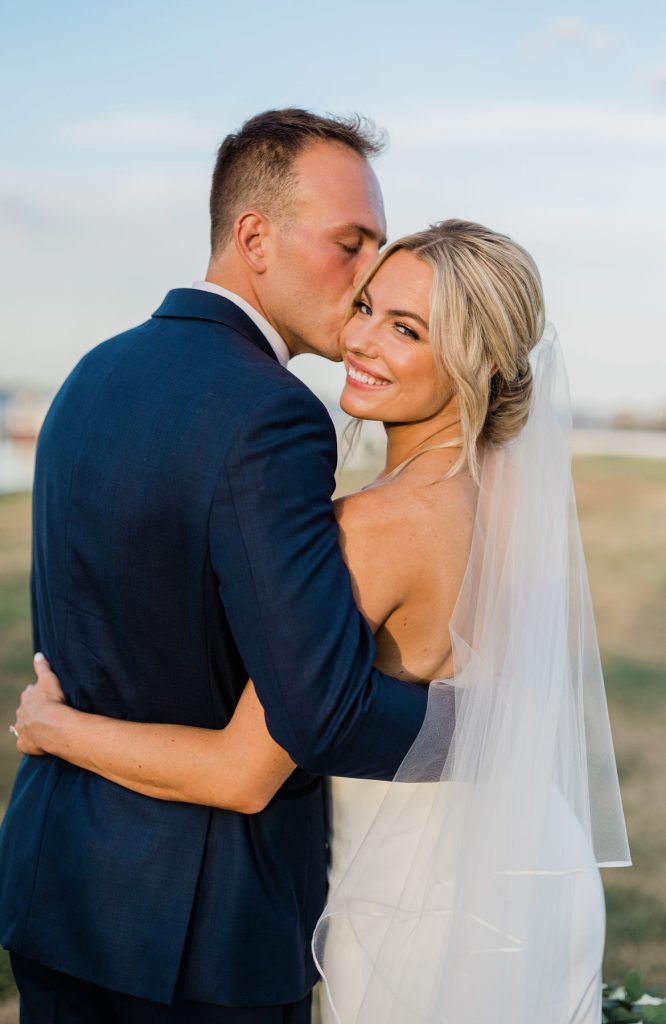 Bride with elegant updo and veil smiles at camera while groom in navy suit kisses her temple at outdoor venue