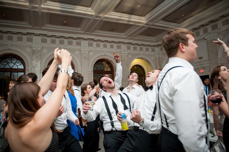 Wedding guests dancing and celebrating energetically in an elegant ballroom with ornate ceiling and arched windows