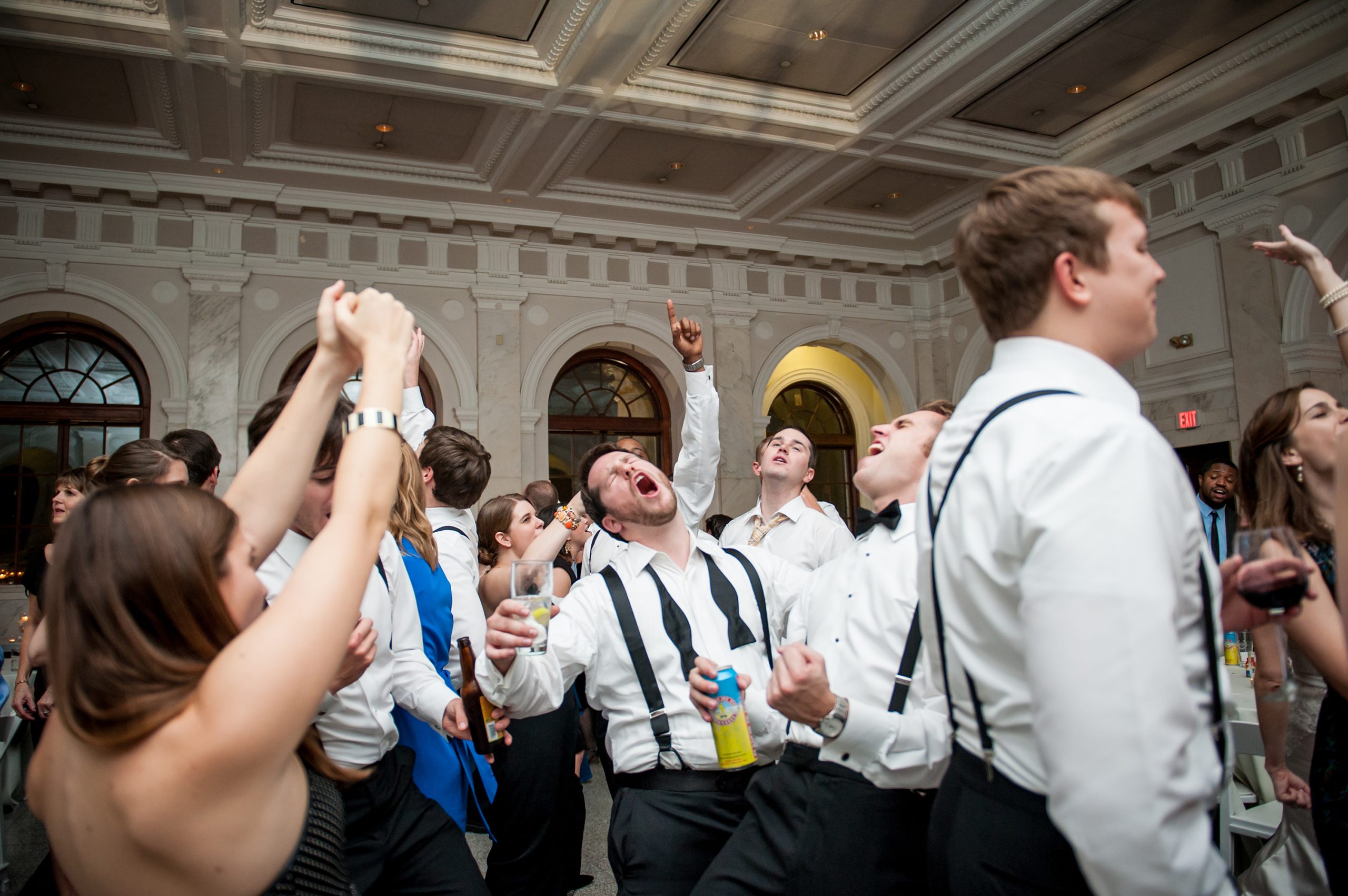 Wedding guests dancing and celebrating energetically in an elegant ballroom with ornate ceiling and arched windows