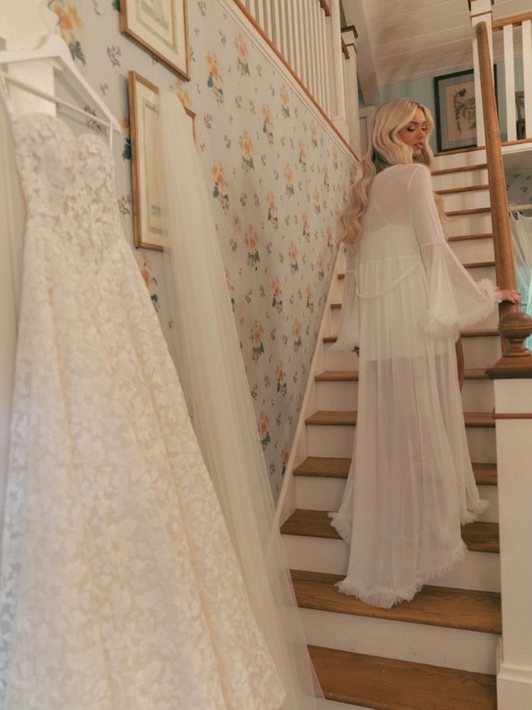Bride in flowing white robe on staircase with lace wedding gown hanging nearby