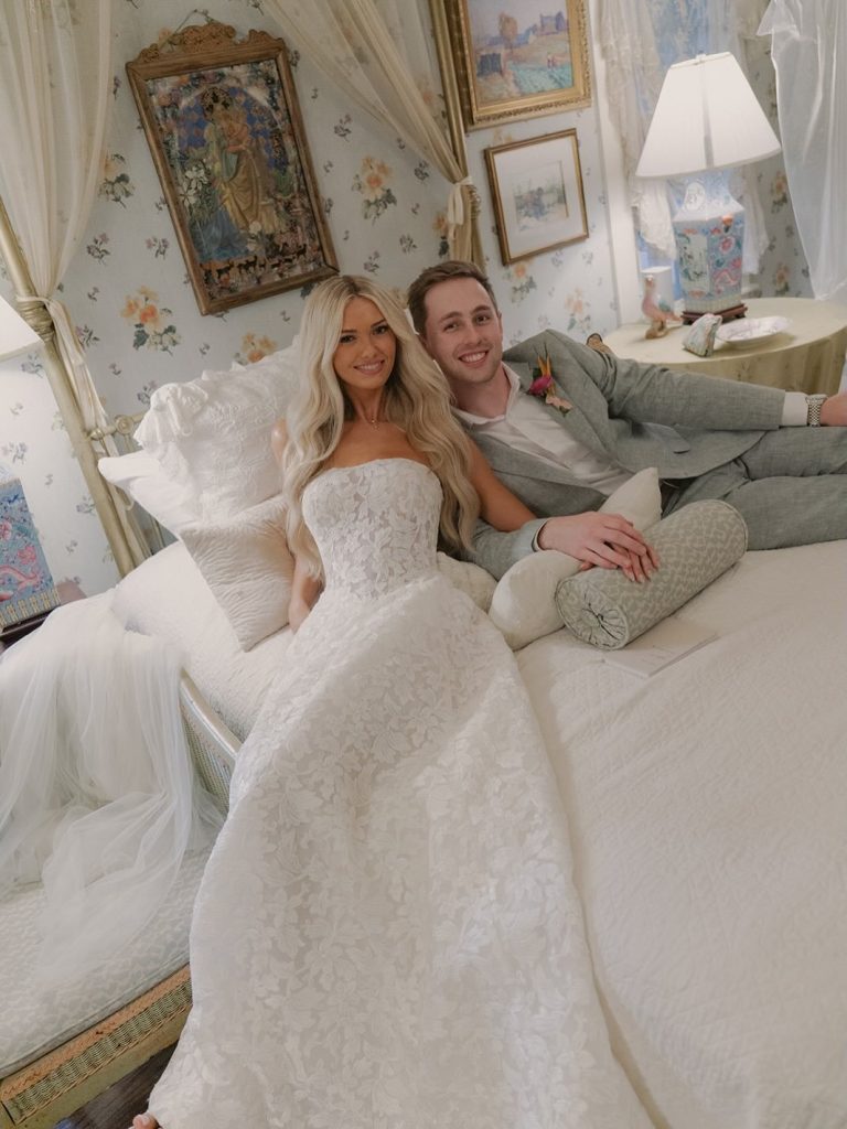 Bride in lace ballgown and groom in gray suit relaxing on bed in elegant room