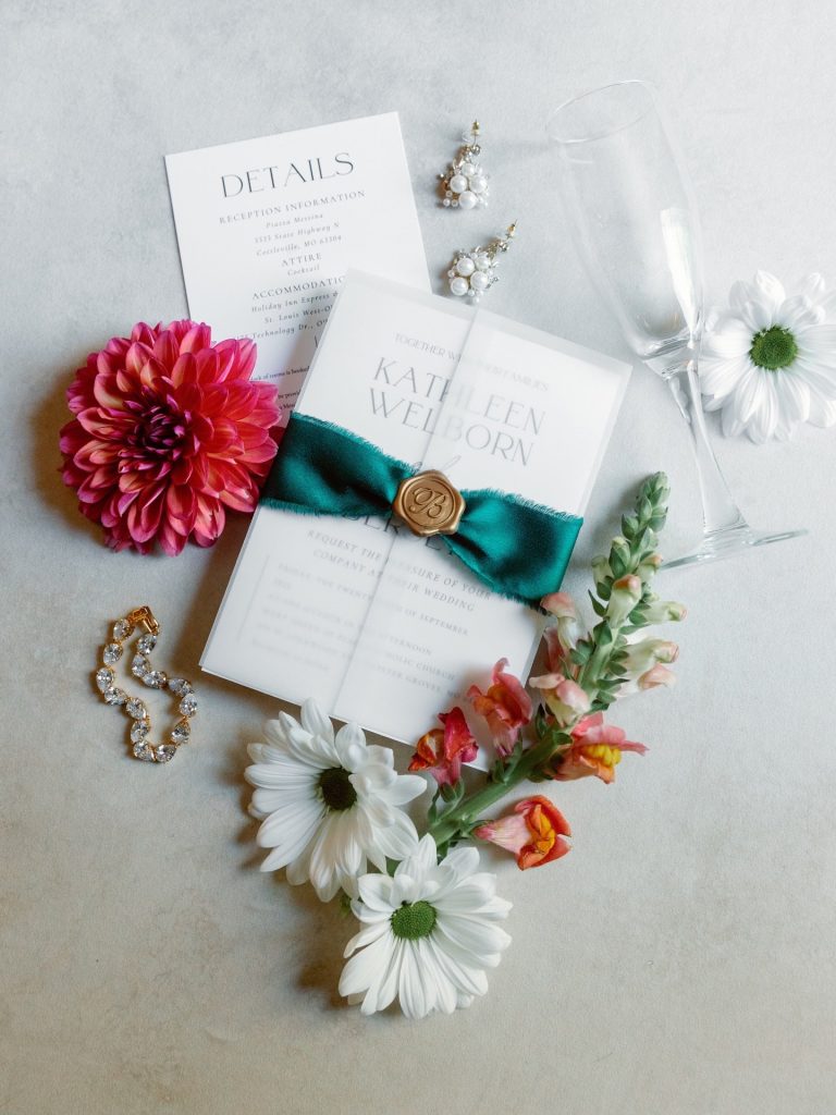 Wedding invitation suite with teal ribbon and wax seal, surrounded by pink dahlia, white daisies, pearl earrings, and crystal bracelet