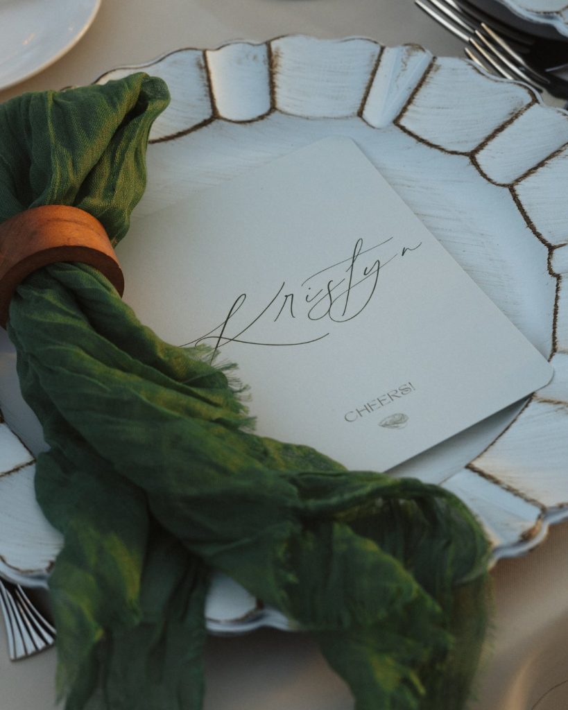 Reception place setting with handwritten escort card, green linen napkin, and scalloped plate at intimate wedding at Addy Sea in Bethany Beach, Delaware.