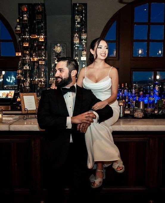 Bride in elegant satin gown sitting on bar counter beside groom in black tuxedo at dimly lit venue