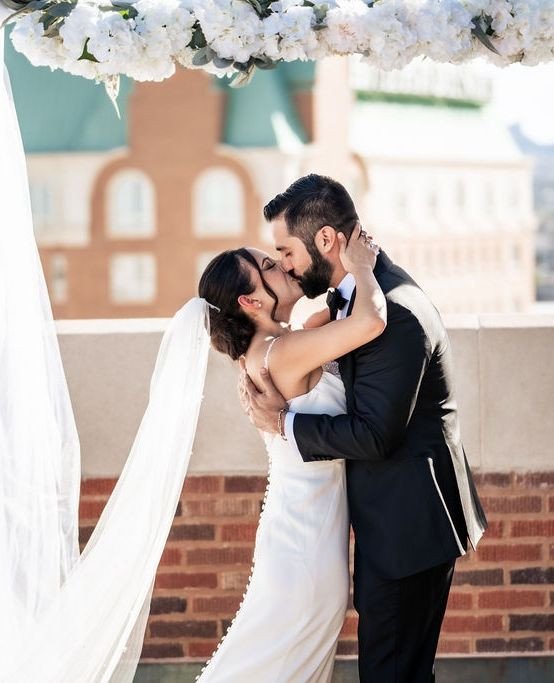 Ali and Joey share a romantic kiss under white floral arbor on rooftop terrace with El Paso cityscape behind them