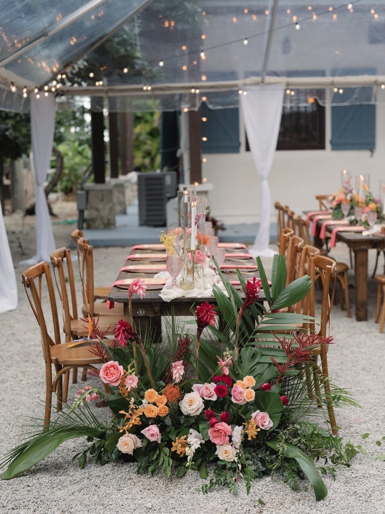Tropical reception tablescape with vibrant floral arrangements, wooden chairs, and string lights under clear tent
