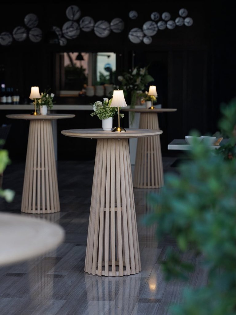 Elegant cocktail tables with pleated bases, gold lamps, and white floral arrangements under string lights at evening reception