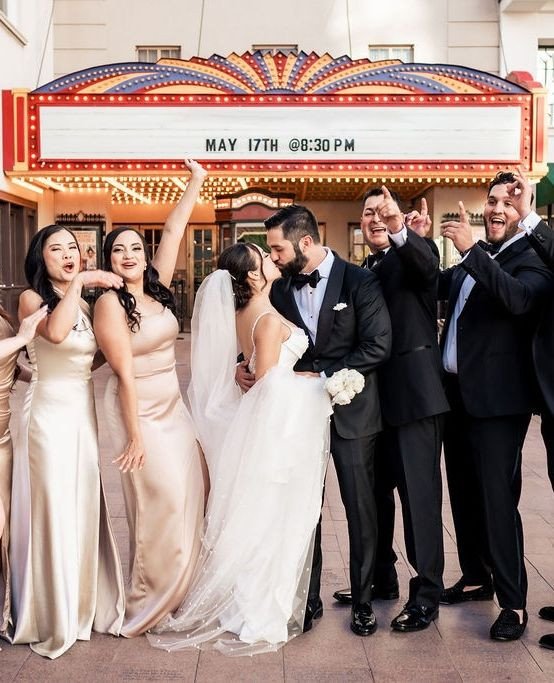 Elegant wedding party portrait with bride and groom kissing beneath historic theater marquee