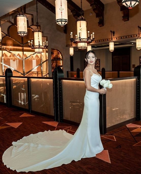 Bride Ali poses in fitted white gown with cathedral train in elegant hotel lobby featuring ornate Spanish-style chandeliers
