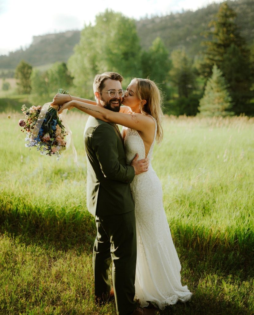 Mallory kisses Andrew's cheek in mountain meadow with wildflower bouquet and evergreen trees in background