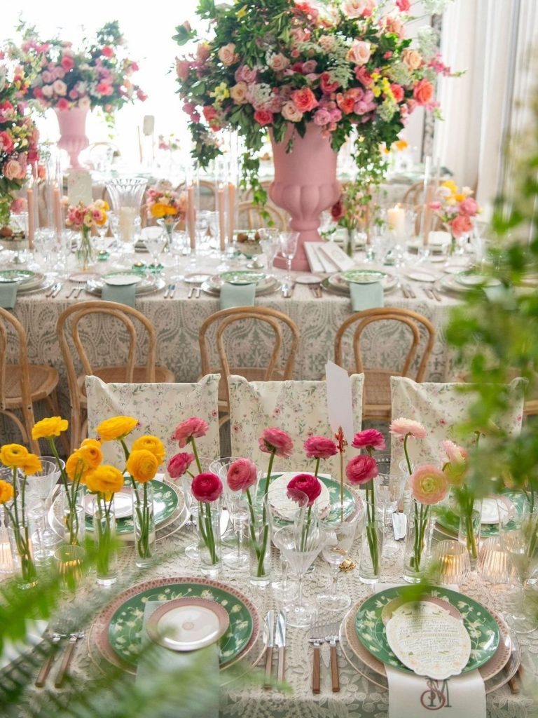 Reception table with pink urn centerpiece, colorful blooms in bud vases, floral-patterned chairs, and green china place settings