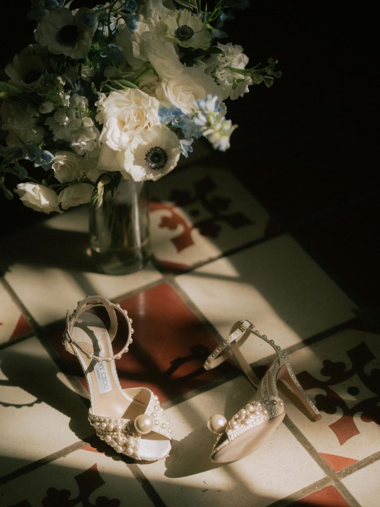 Pearl-embellished bridal heels and wedding rings beside bouquet of white blooms in dramatic window light