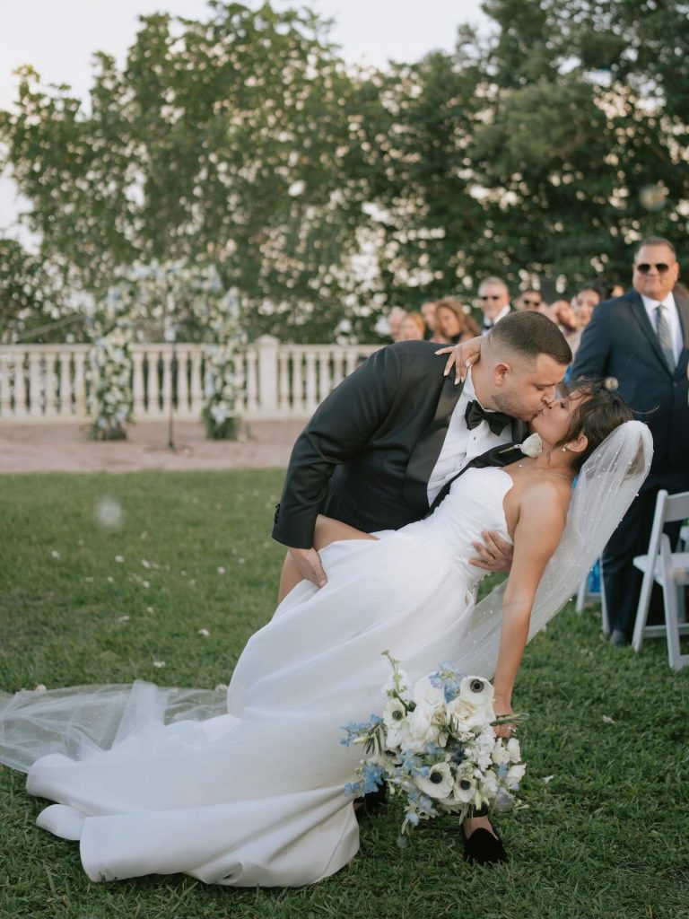 Newlyweds share romantic dip kiss on lawn after outdoor ceremony with guests watching