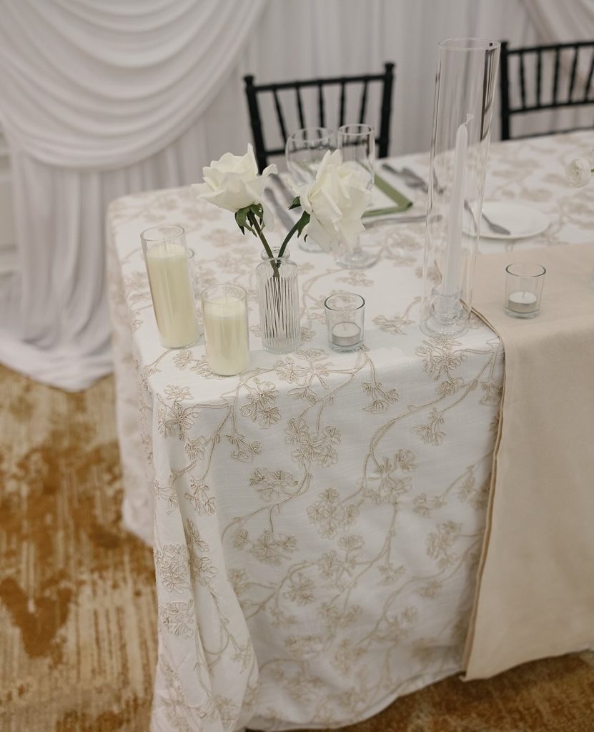 Elegant sweetheart table with white flowers in crystal vase, pillar candles, and embroidered linen tablecloth with black chiavari chairs