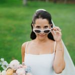 Bride in modern white gown and sunglasses holding pastel bouquet on lawn