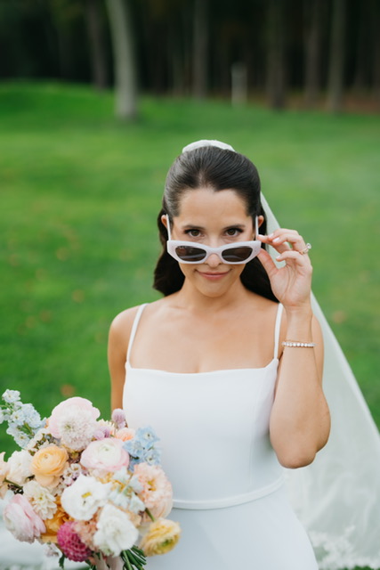 Bride in modern white gown and sunglasses holding pastel bouquet on lawn