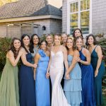 Bridal party in mismatched blue and green dresses gathered outside gray house