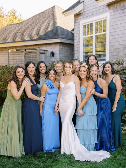 Bridal party in mismatched blue and green dresses gathered outside gray house