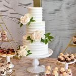White tiered wedding cake adorned with fresh white roses on a cake stand with dessert table in background